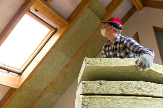A builder wearing a hard hat, safety glasses, and gloves is fitting insulation in a loft conversion.